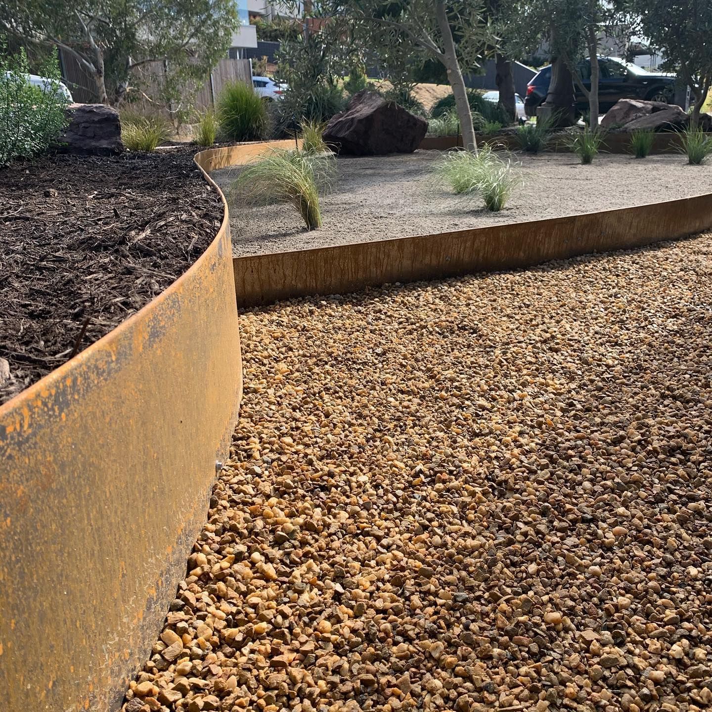A curved rusted metal border separates dark soil and gravel path in a garden with plants and trees.
