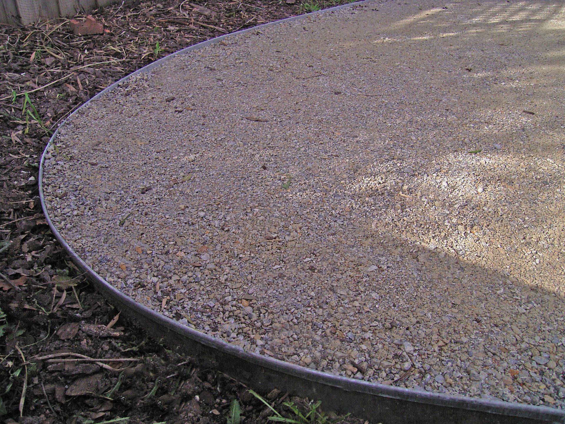 Gravel pathway bordered by a dark metal edge, curving towards the viewer. Brown and beige tones, outdoor setting.