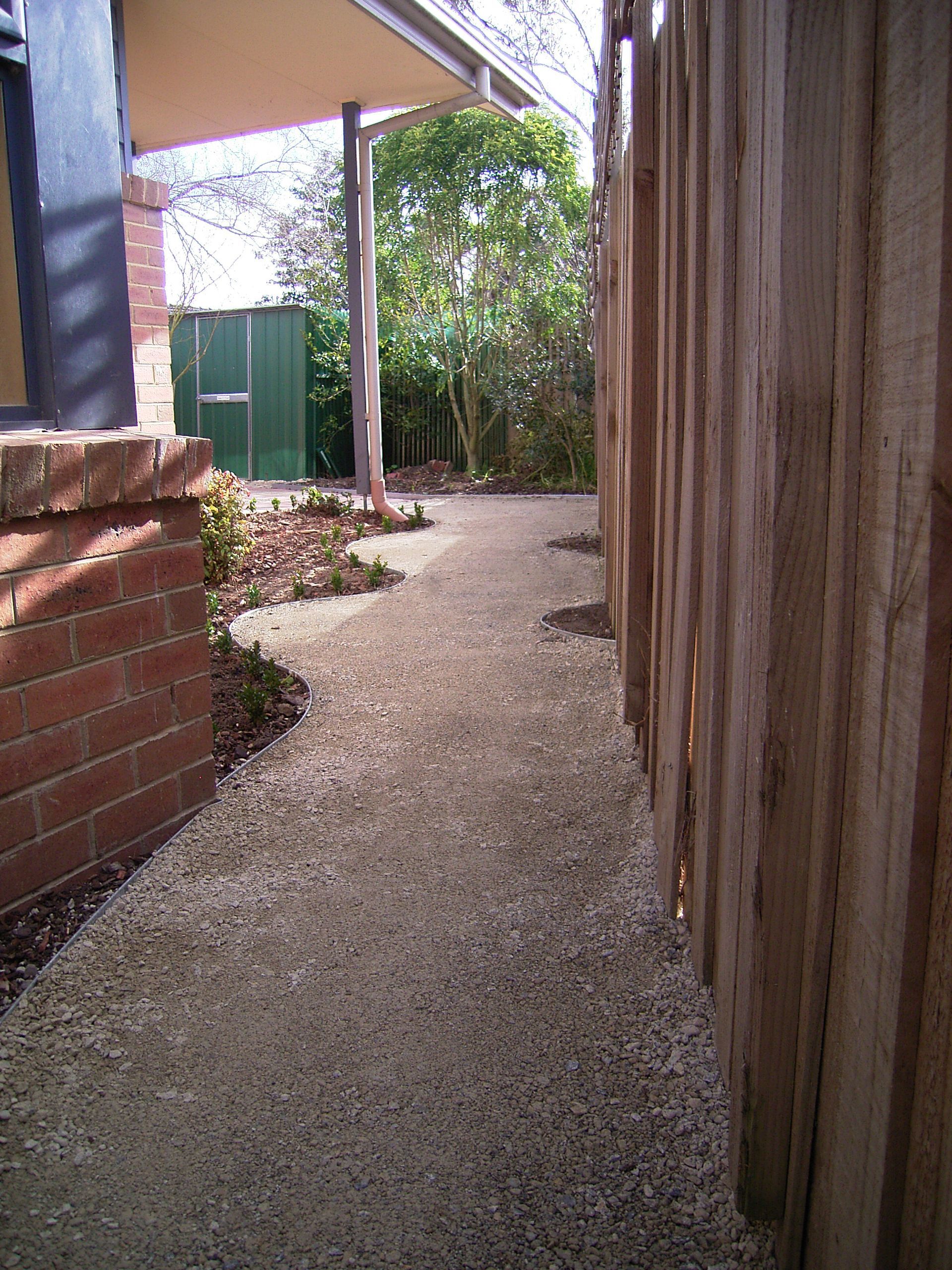 Gravel path alongside a wooden fence, leading towards a green shed and trees; brick wall on the left.