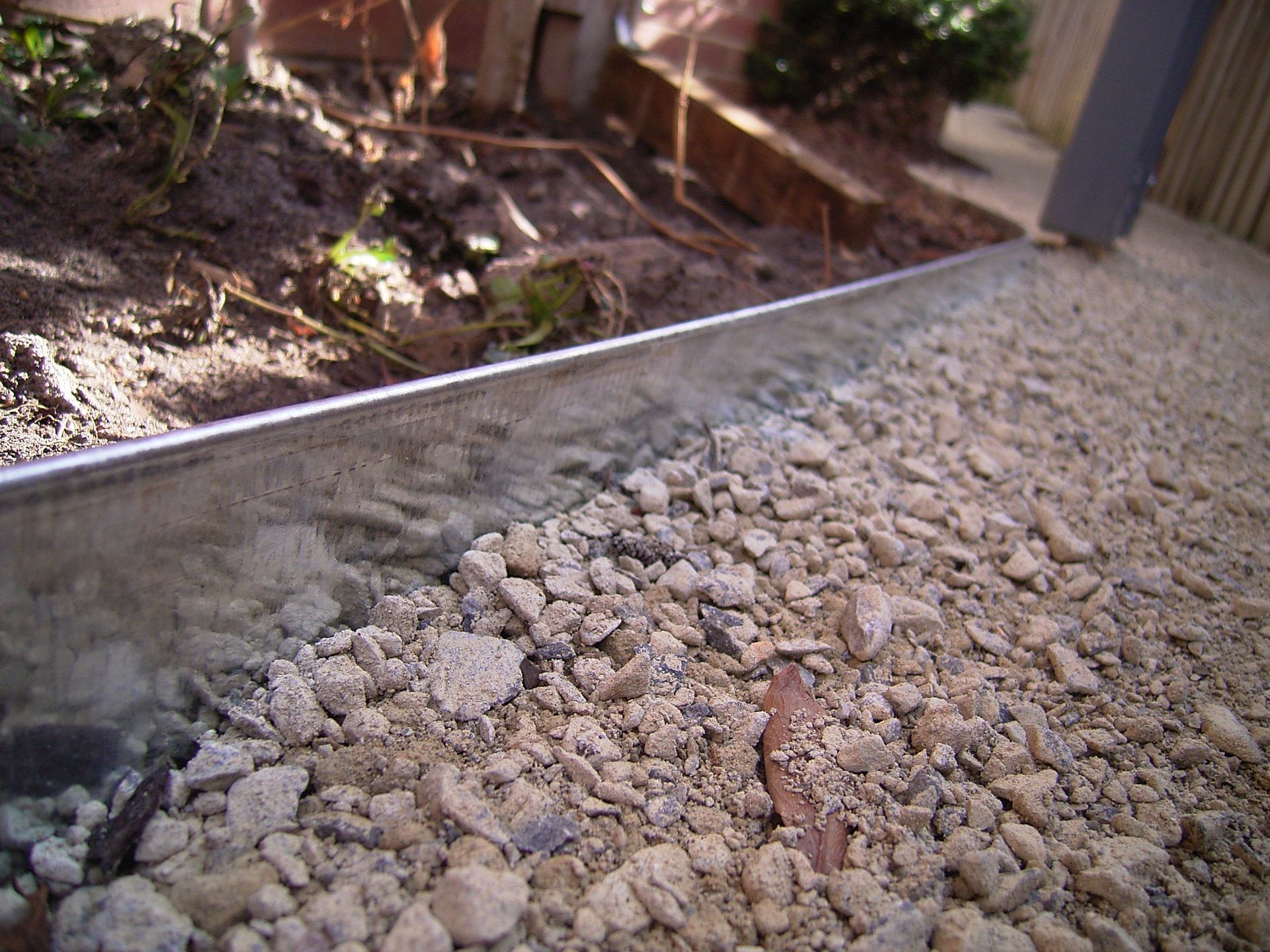 Close-up of gravel edge next to soil bed, separated by a metal barrier in sunlight.