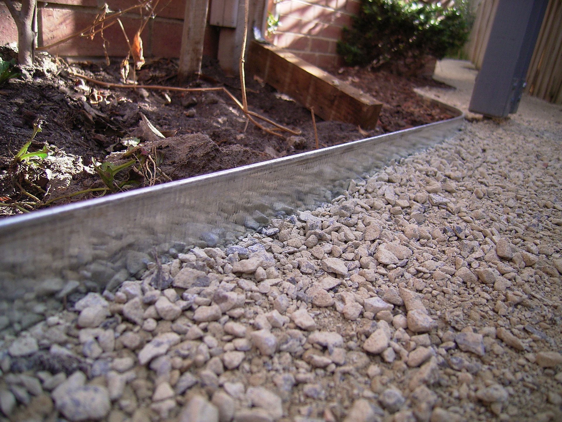 Gravel path edge with metal edging, adjacent to a garden bed with soil and small plants.