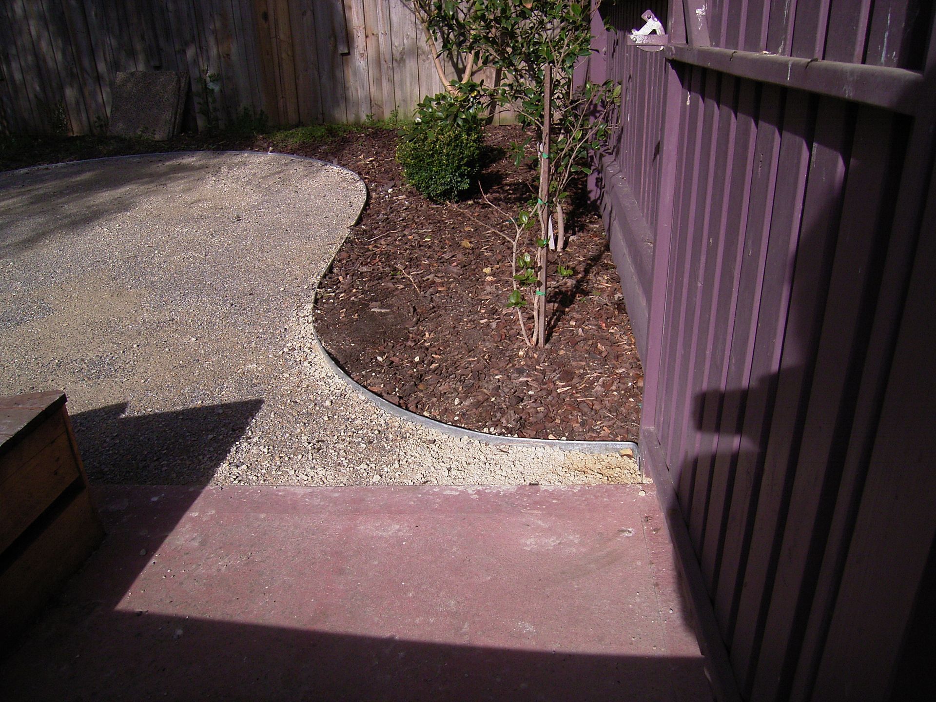 A gravel path curves around a mulched garden bed next to a purple wooden fence.