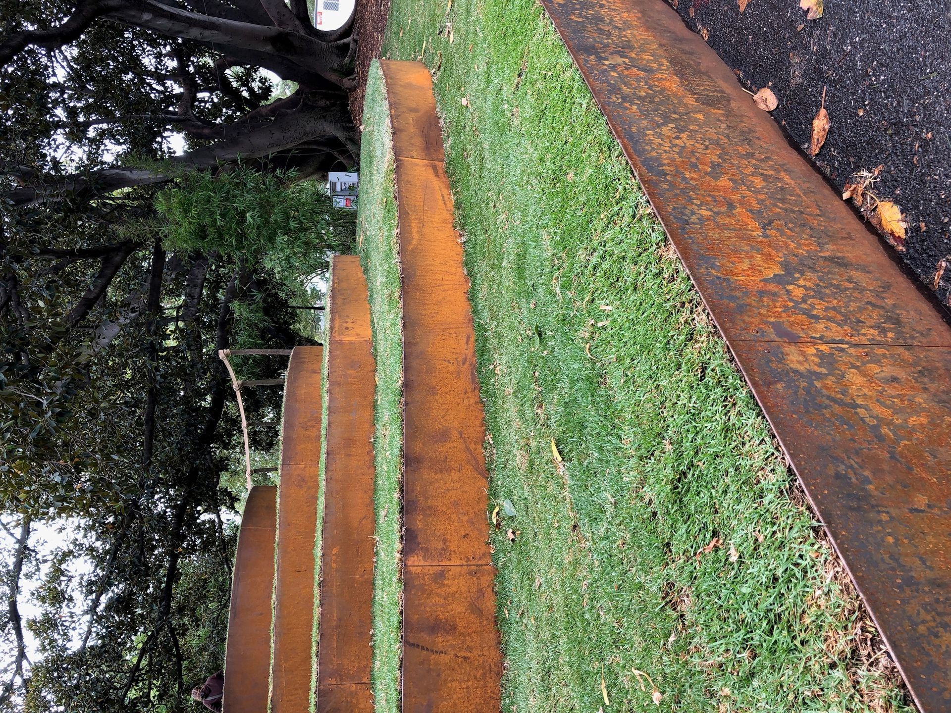 Rust-colored steel sculpture curving along a grassy area, alongside asphalt.