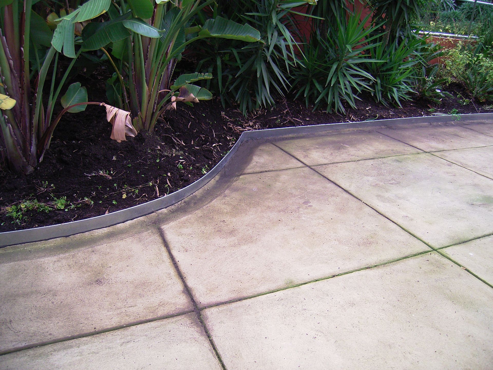 Stone patio edge with metal edging bordering a dark soil garden bed with greenery.