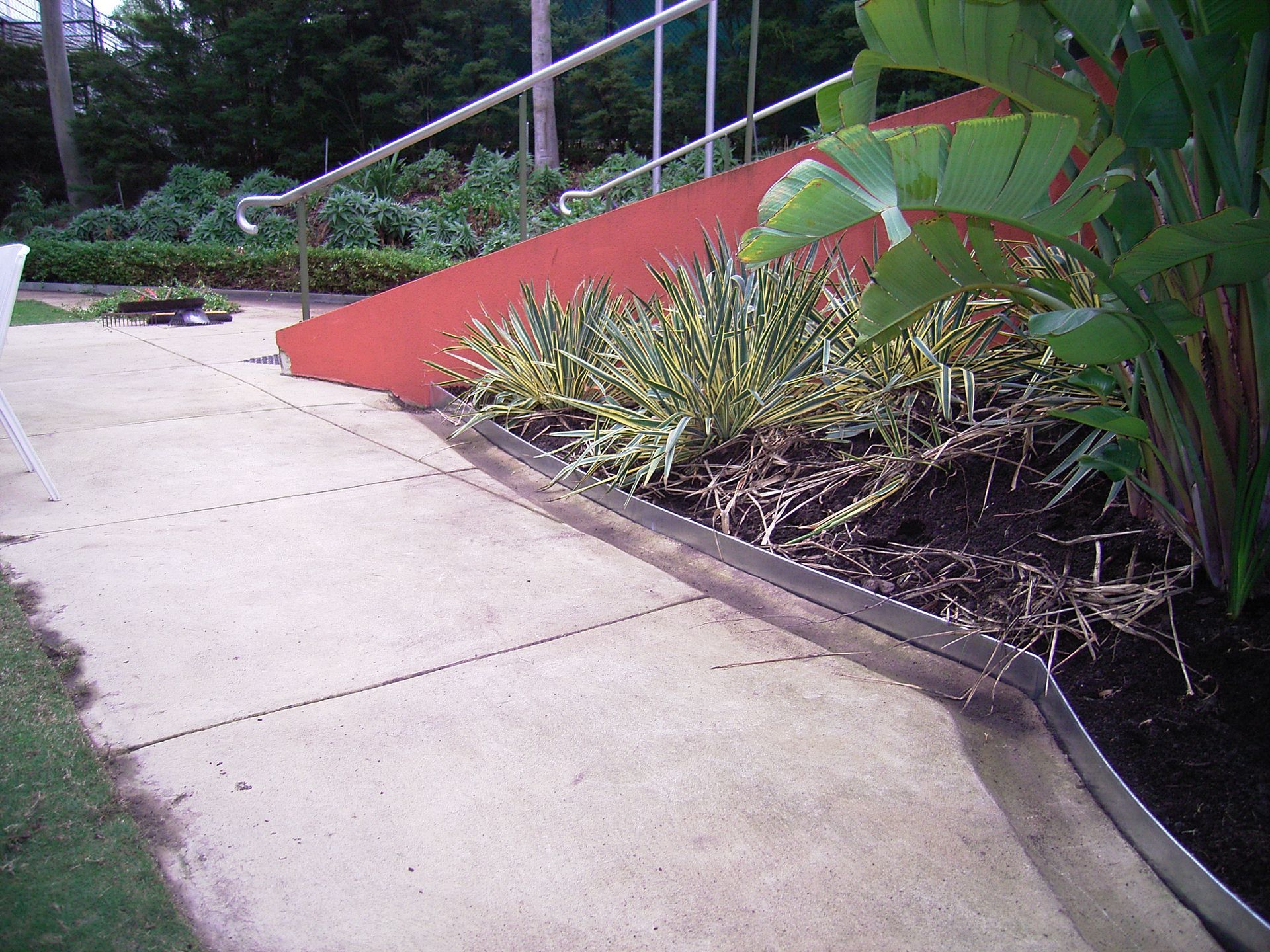 Concrete pathway next to a garden bed, with red ramp and metal railing in background.