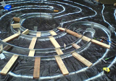 Metal spiral being constructed with wood supports on a black surface marked with white lines.