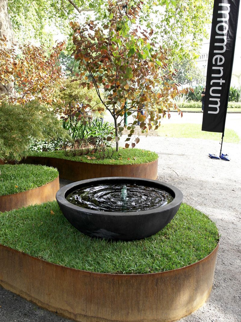 A modern outdoor water feature with a dark bowl fountain, surrounded by green grass and rust-colored metal edging.