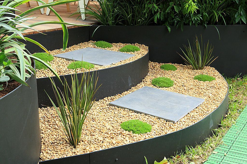 Curved, tiered garden bed with stone steps, gravel, and small plants. Dark border.