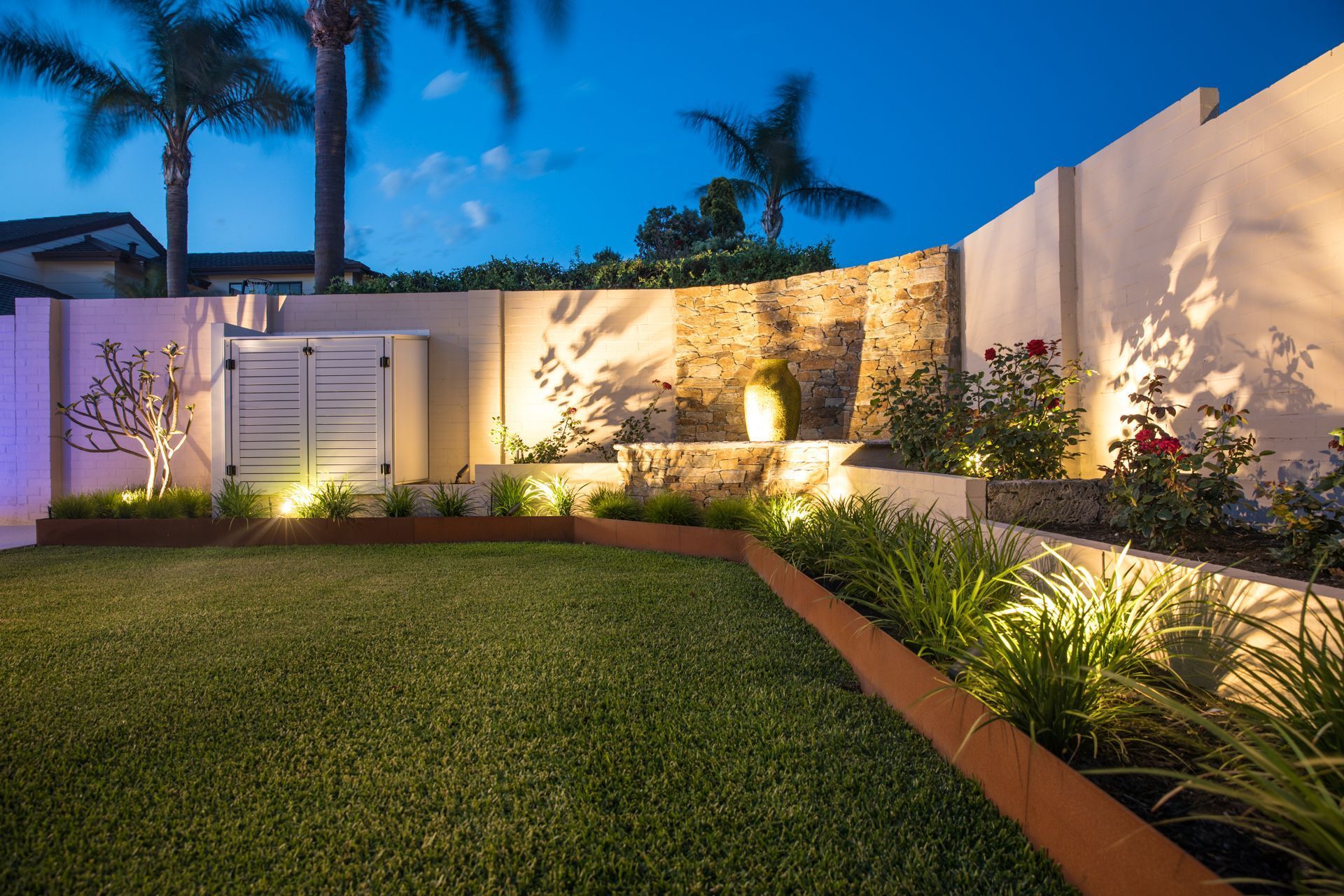Well-lit backyard with lawn, garden beds, and decorative stone wall. Nighttime with warm lighting.