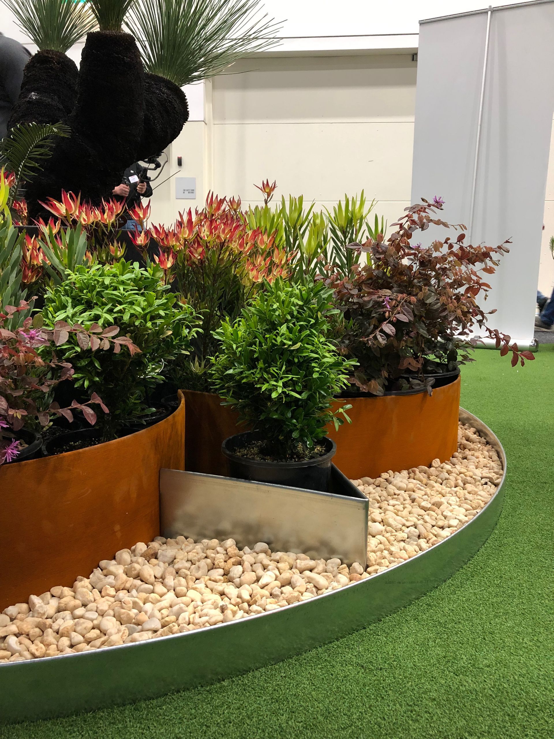A garden display with various plants in rusty metal containers on a bed of beige pebbles, set on green turf.