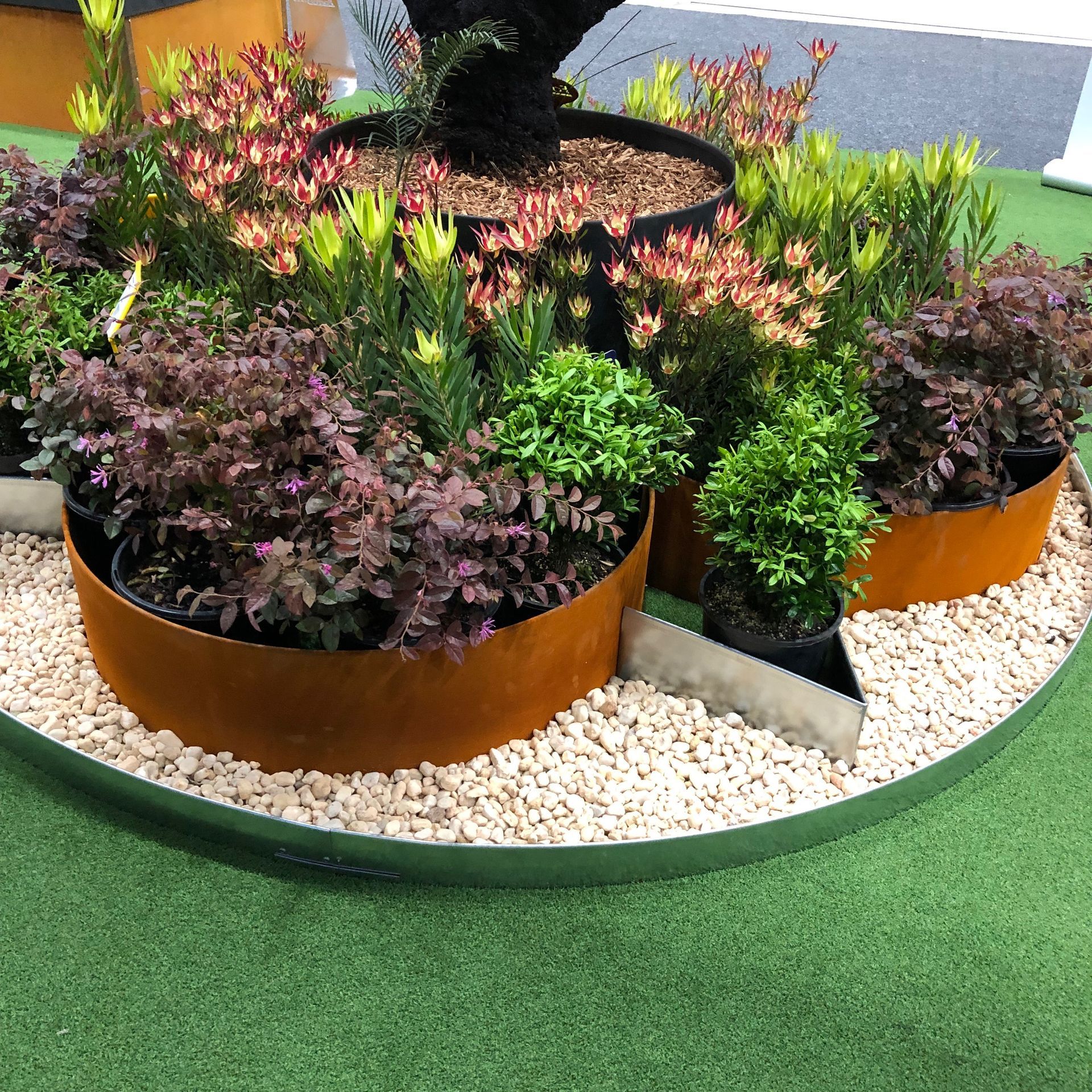 Colorful plants in rusty metal planter beds on gravel, displayed on green turf.