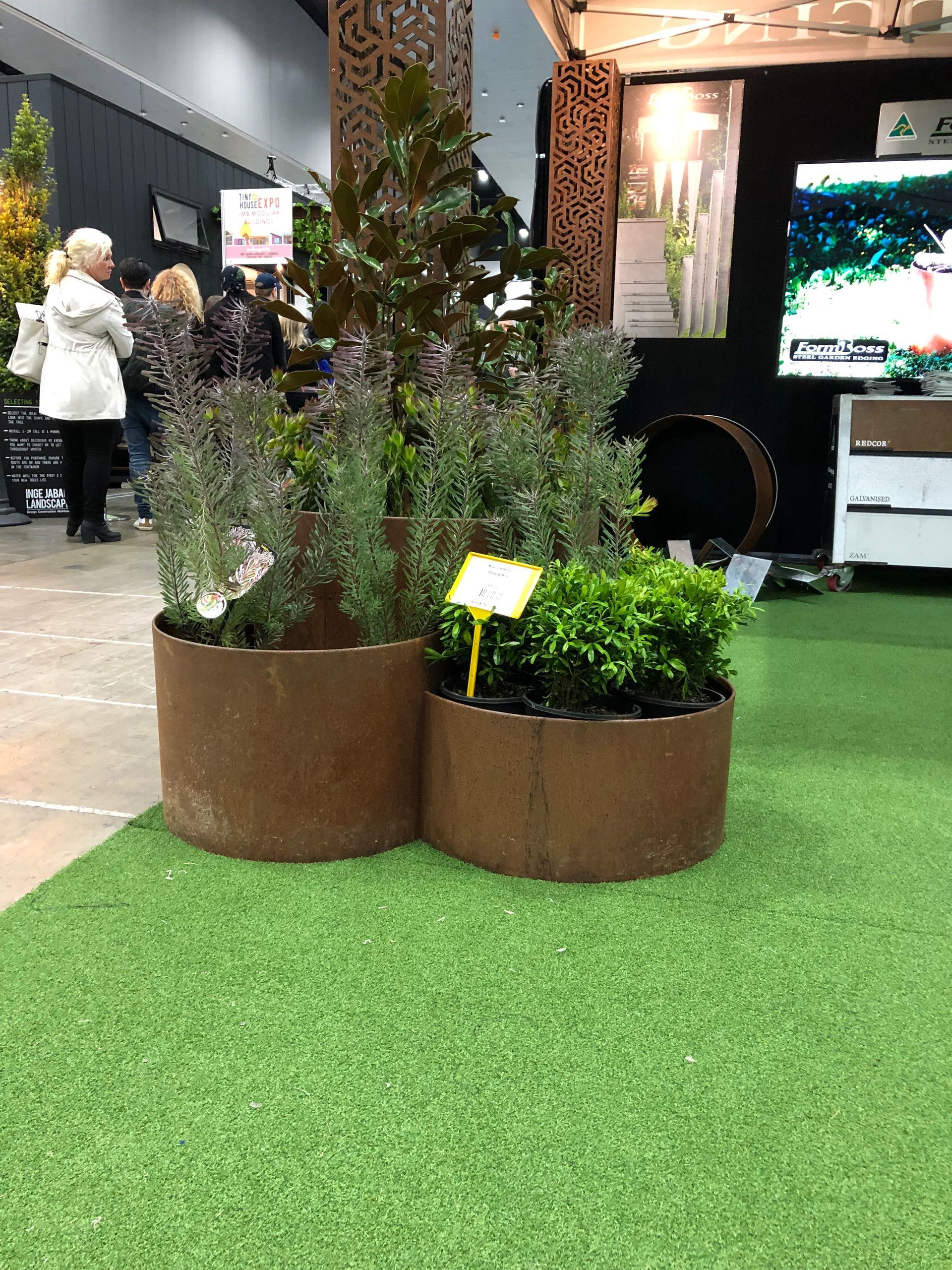Three rusty metal planters with green plants on a green, artificial lawn. People in the background.