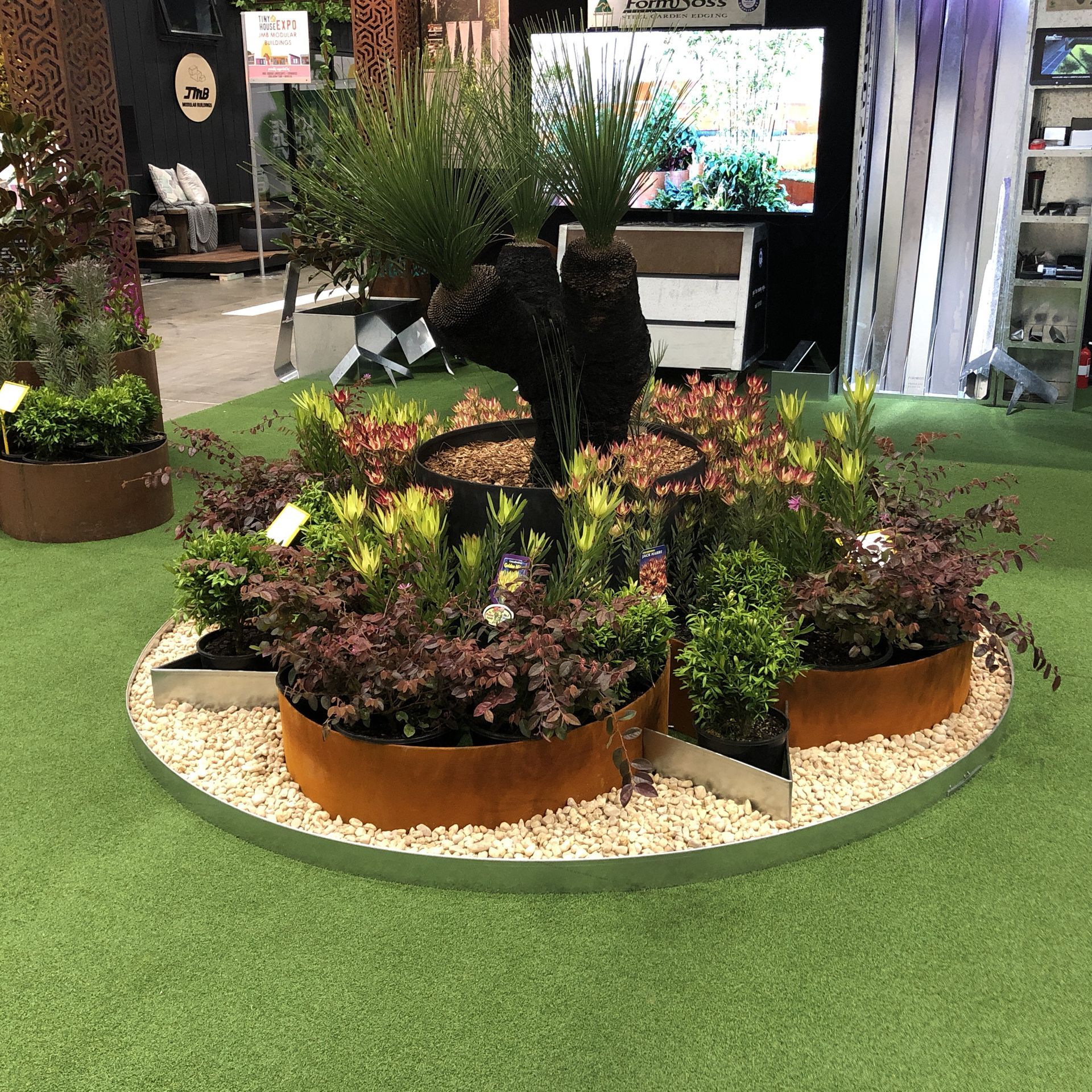Display of plants in a circular bed with rusted metal edging on a green surface.