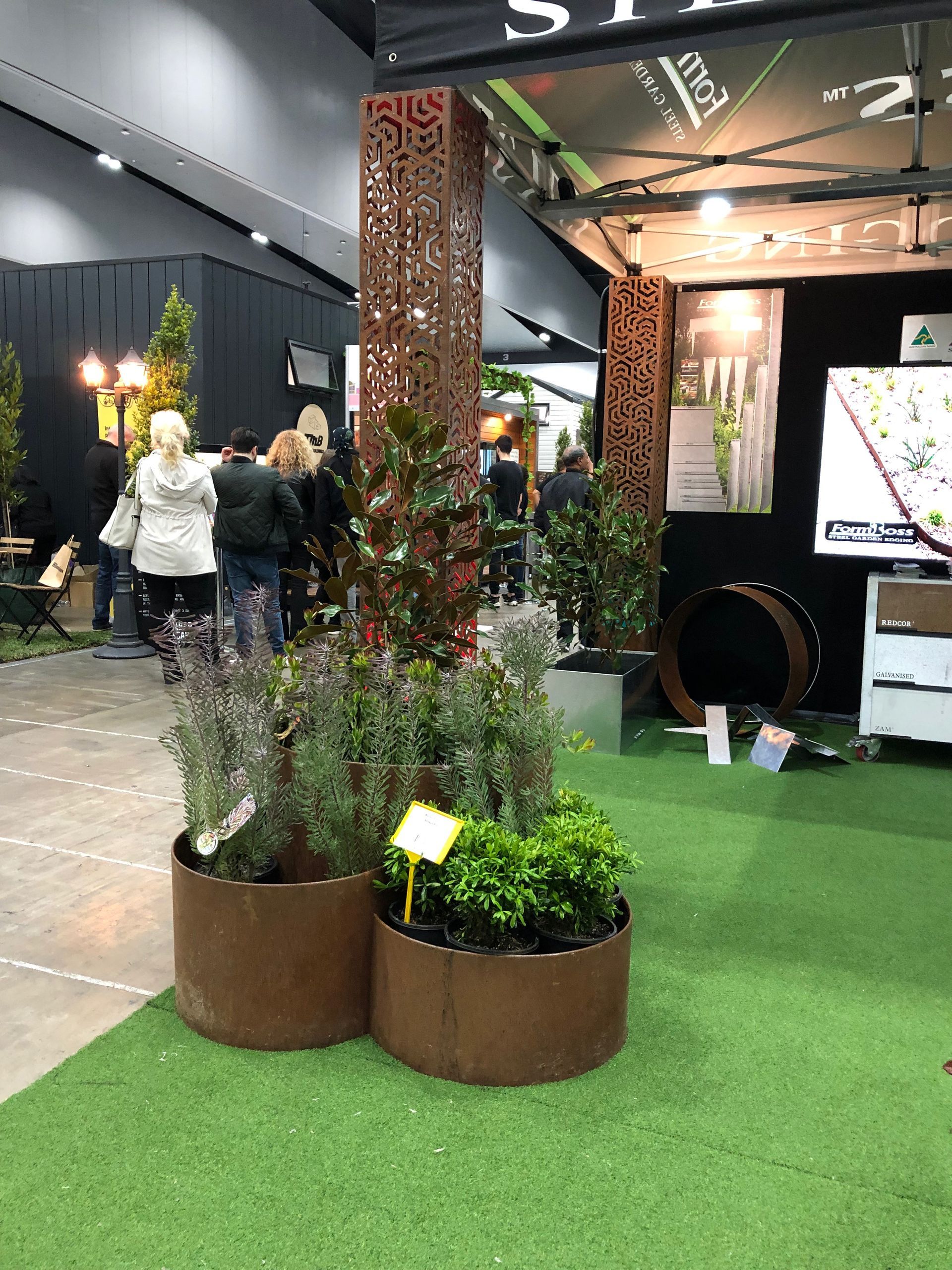 A trade show booth with potted plants, metal columns, and a green turf floor.