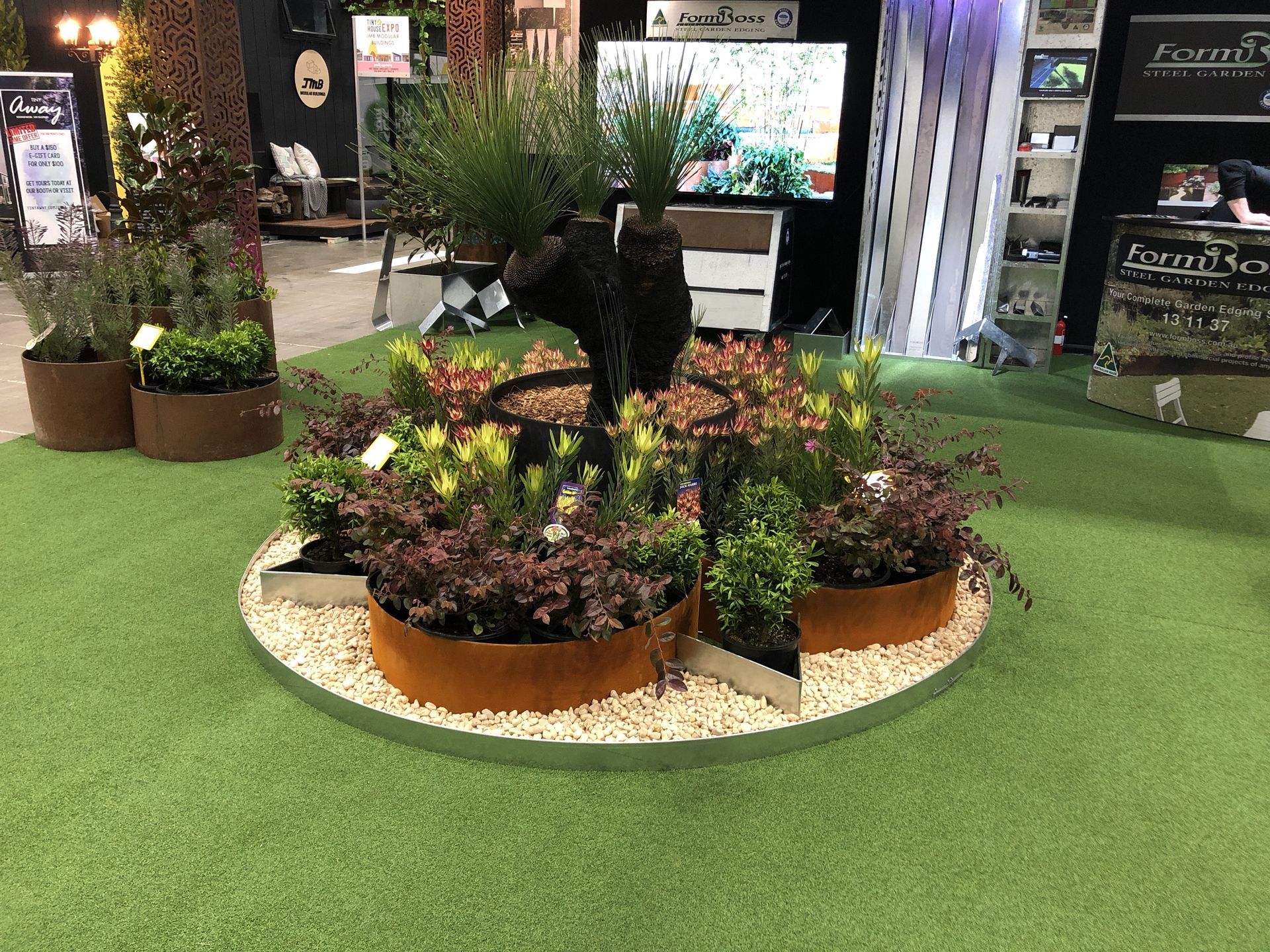 A display of potted plants in a circular arrangement, surrounded by white pebbles, set on green turf.