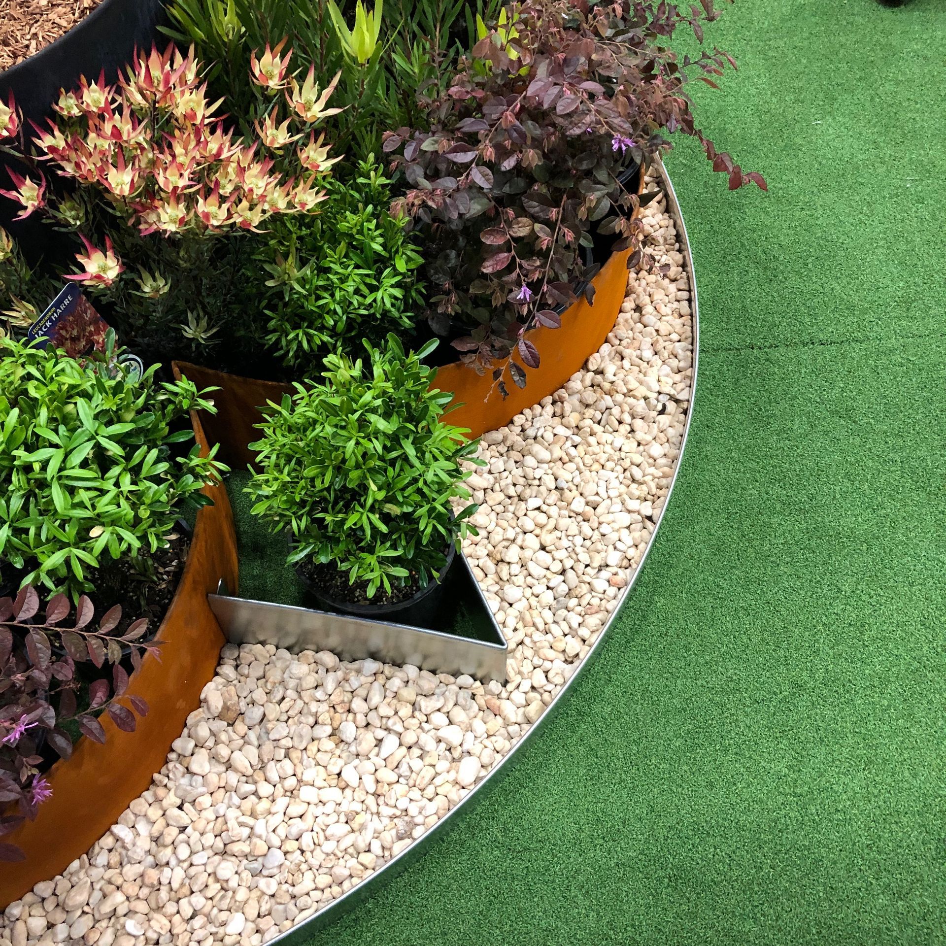 Circular garden bed with vibrant plants, rust-colored edging, and white gravel, adjacent to green turf.