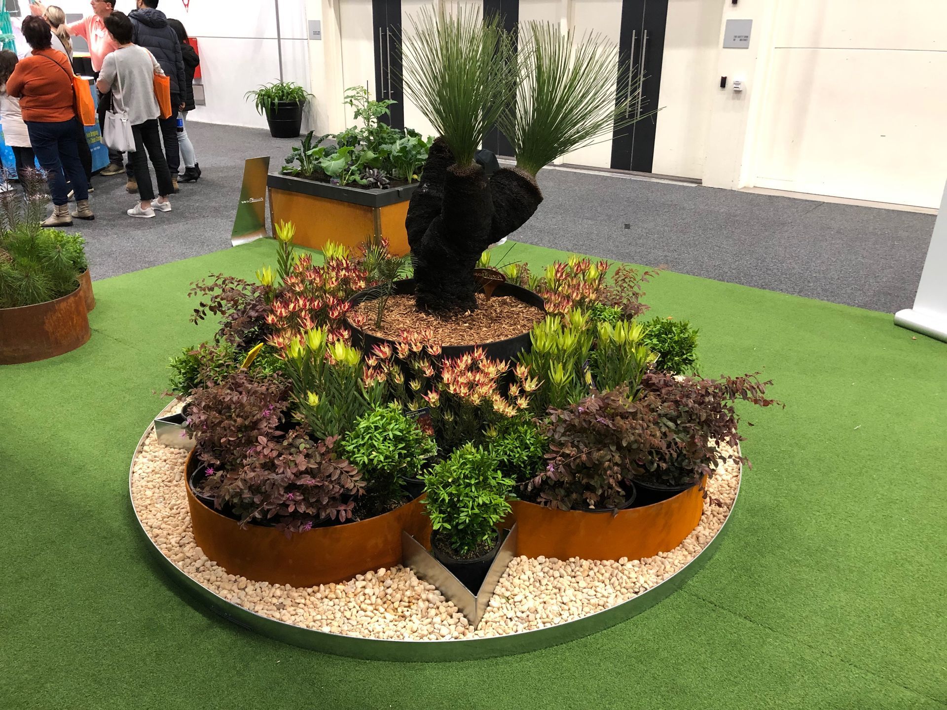 A circular garden display on green turf. A central, dark plant is surrounded by smaller plants in rust-colored planters.