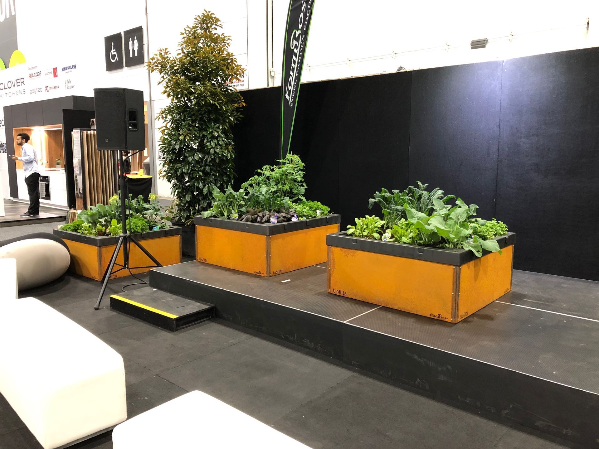 Three orange planters with leafy greens on a black stage. A speaker and trees are in the background.