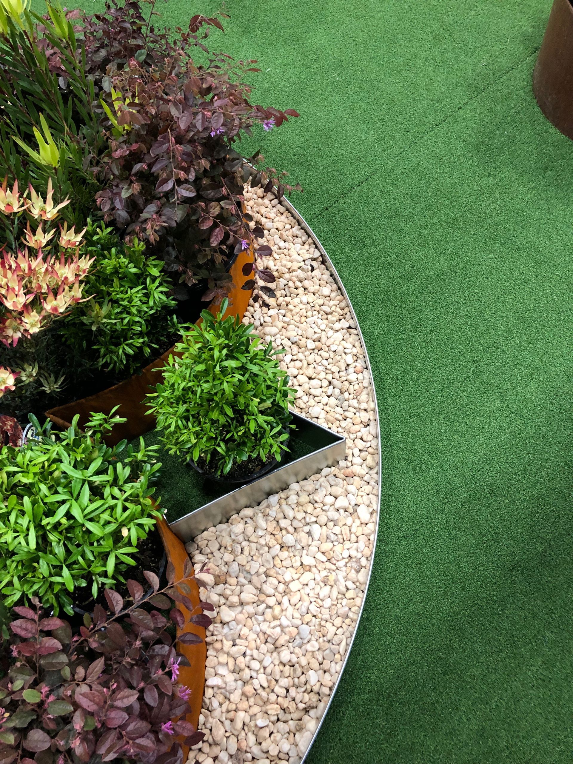 Round garden bed with beige gravel, varied green and purple plants, and artificial green turf.