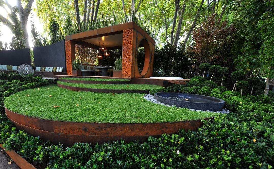 Outdoor garden with rust-colored metal structure, tiered lawn, and dark water feature. Lush green foliage surrounds.