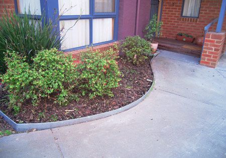 Concrete walkway curves past a landscaped garden bed with green bushes and mulch, beside a brick building.