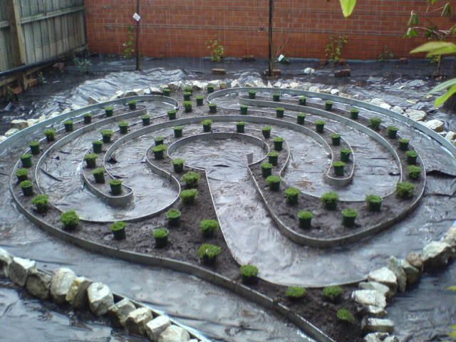 A garden labyrinth with a metal border, containing small plants. Surrounded by a black ground cover and rocks.
