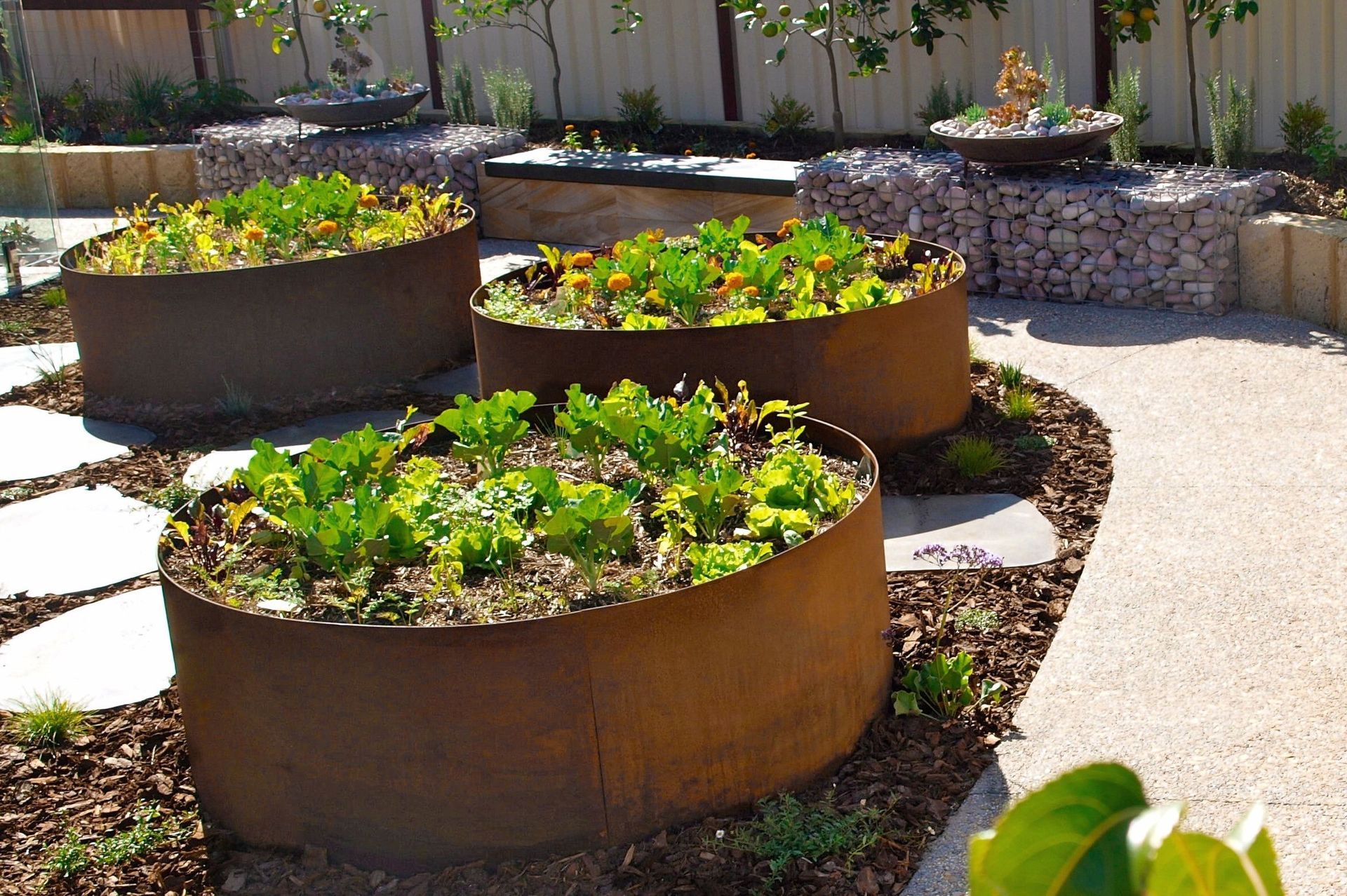 Oval-shaped rusty metal planters filled with green plants in a garden setting, surrounded by stepping stones and decorative rocks.