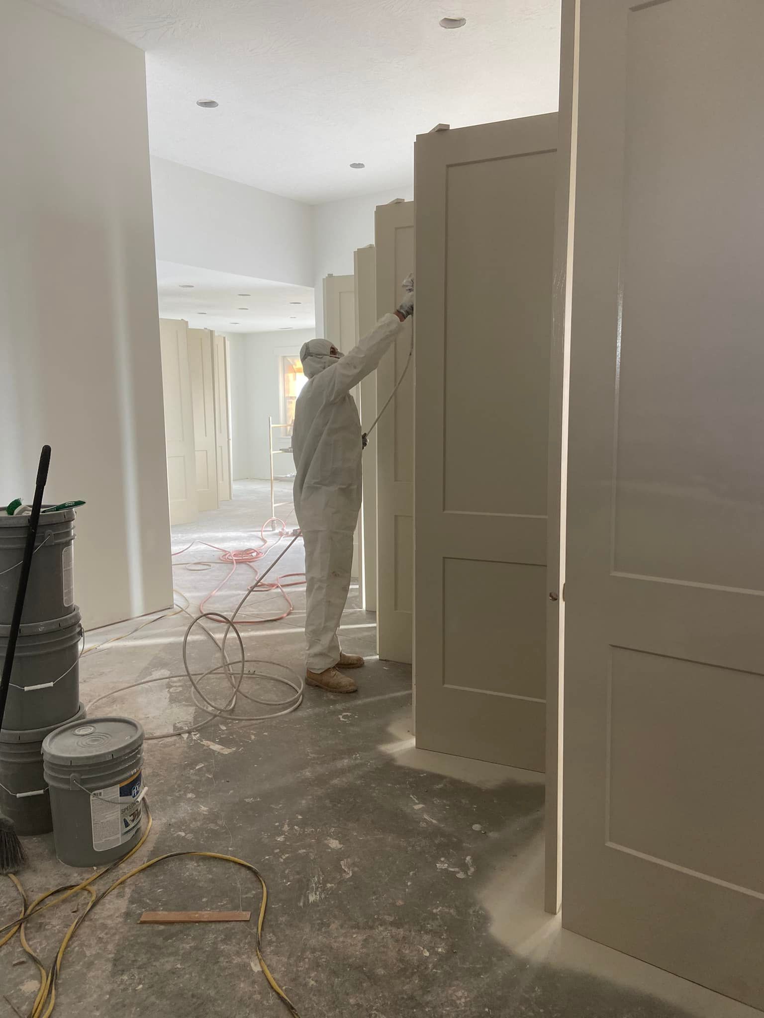 A worker in protective clothing uses a paint sprayer on tall doors in a bright, unfinished room with buckets on the floor.