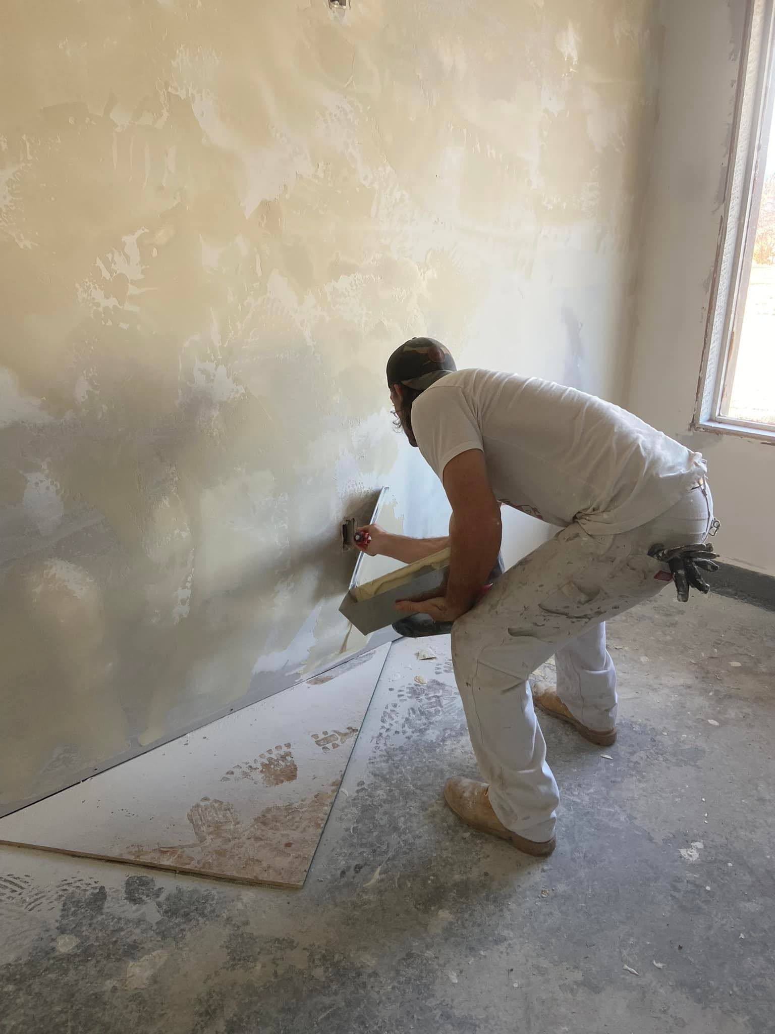 A person in work clothes uses a trowel to apply plaster or joint compound to a textured interior wall.
