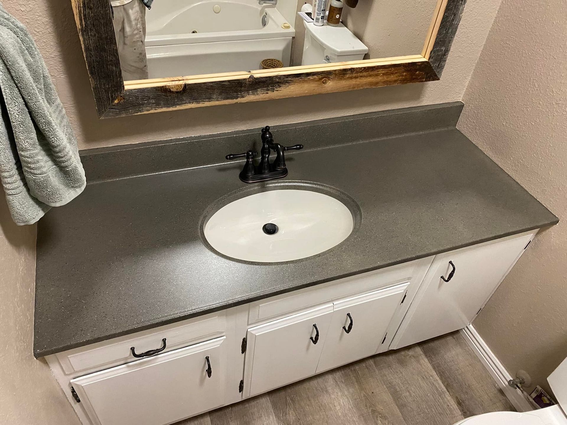A white bathroom vanity with a dark gray speckled countertop, a single oval sink, and a rustic wooden-framed mirror above.