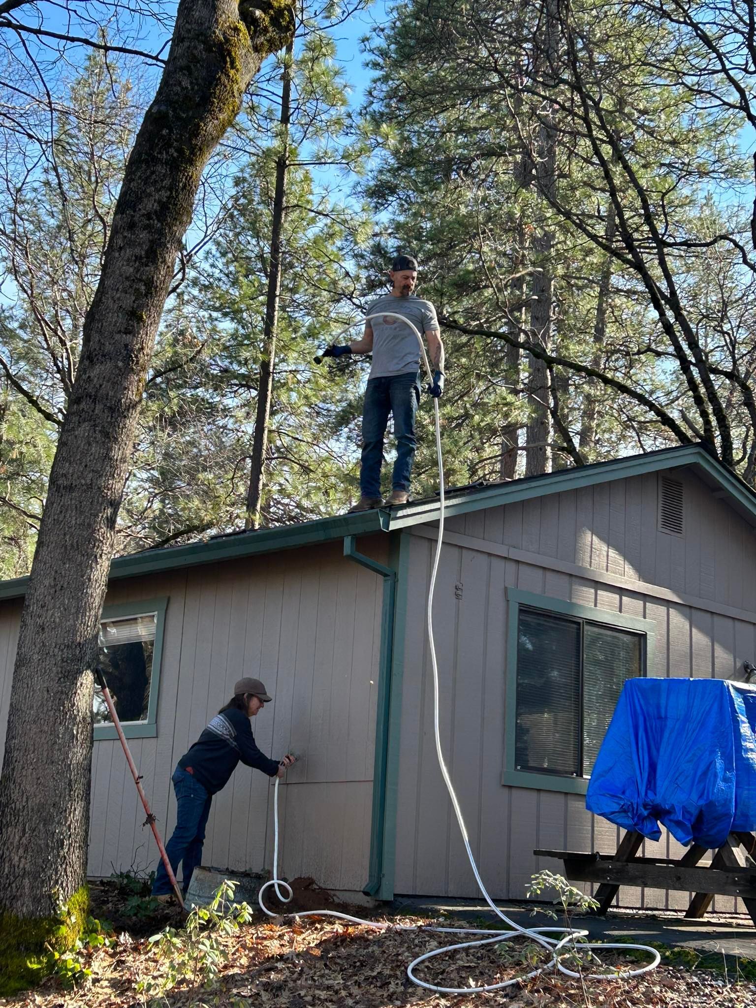 Two people working outside a house; one person stands on the roof holding a rope, while another stands on the ground.