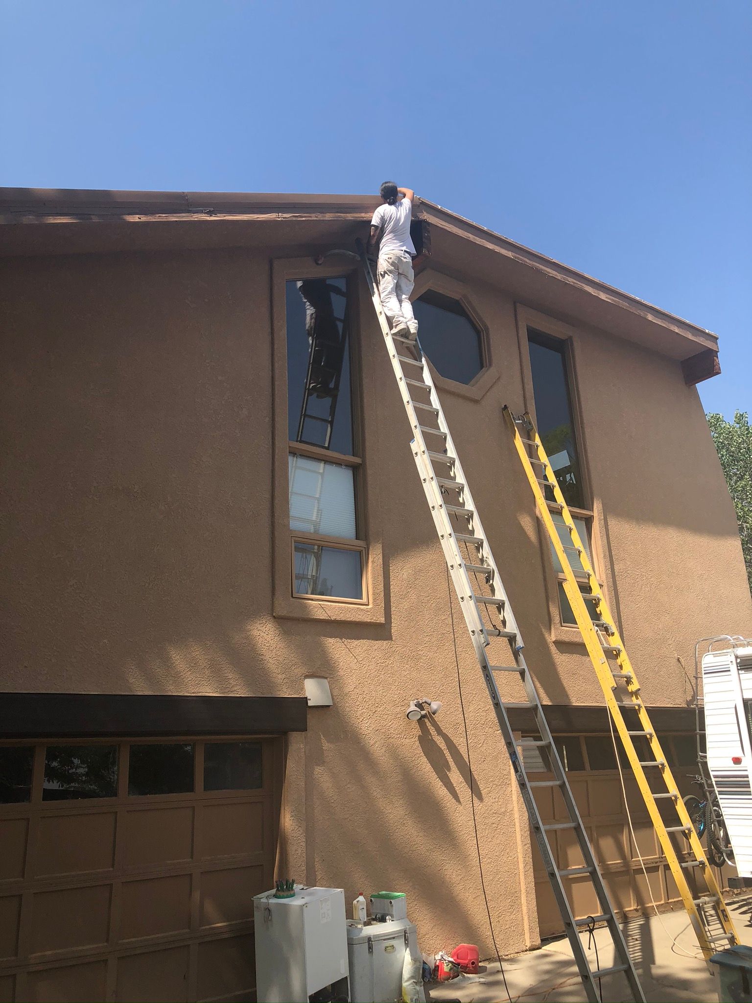 A worker stands on a tall ladder, painting the exterior trim of a multi-story, tan-colored stucco home.