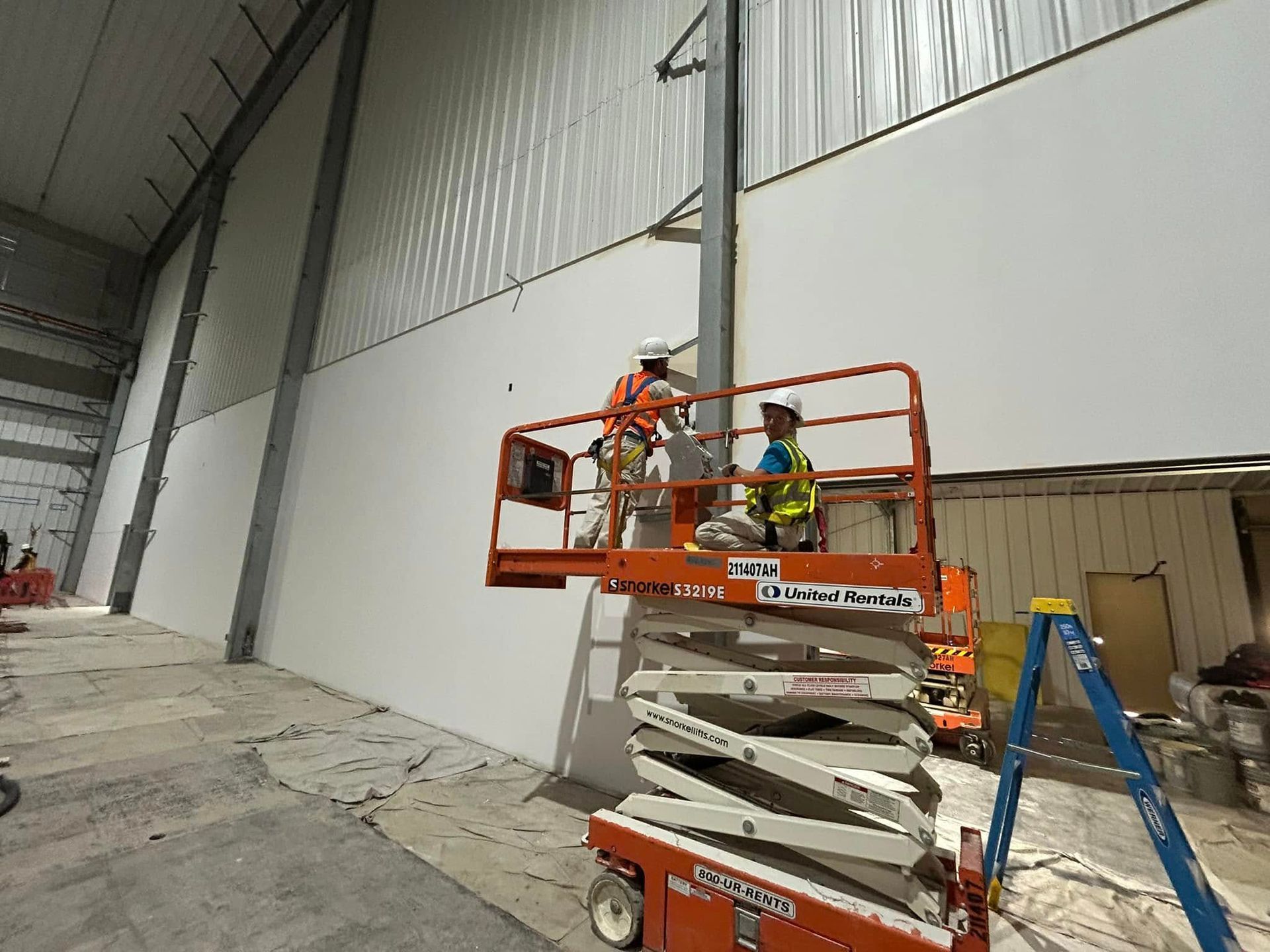 Two workers in hard hats and safety vests stand on an orange scissor lift to paint a large white interior warehouse wall.