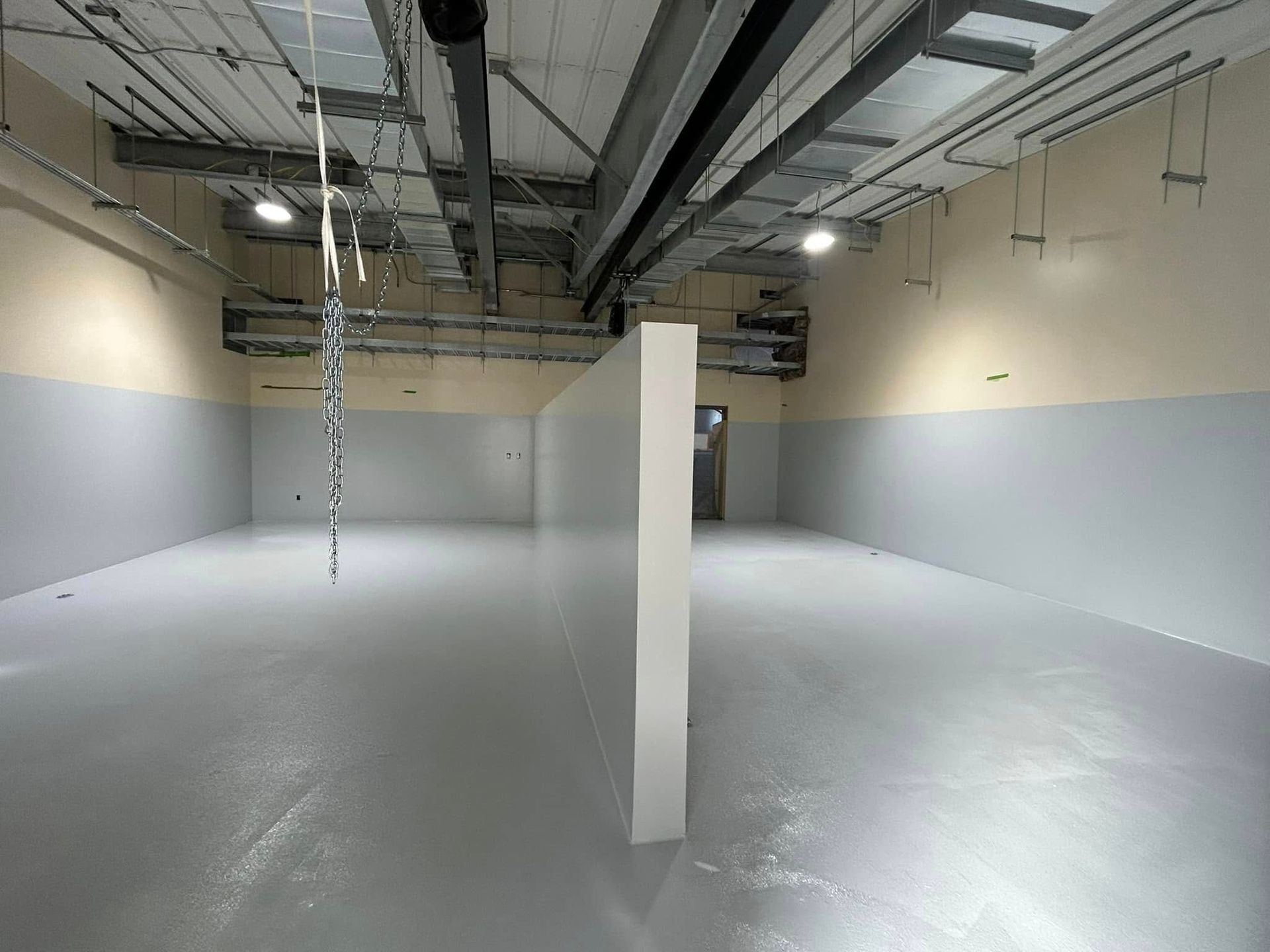 An empty, industrial room with gray walls and flooring, a central partition wall, and exposed overhead ductwork.