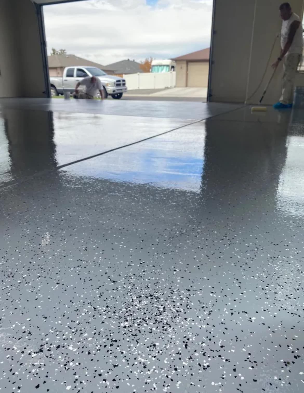 Two workers apply a glossy grey epoxy finish with black and white decorative flecks to a garage floor.