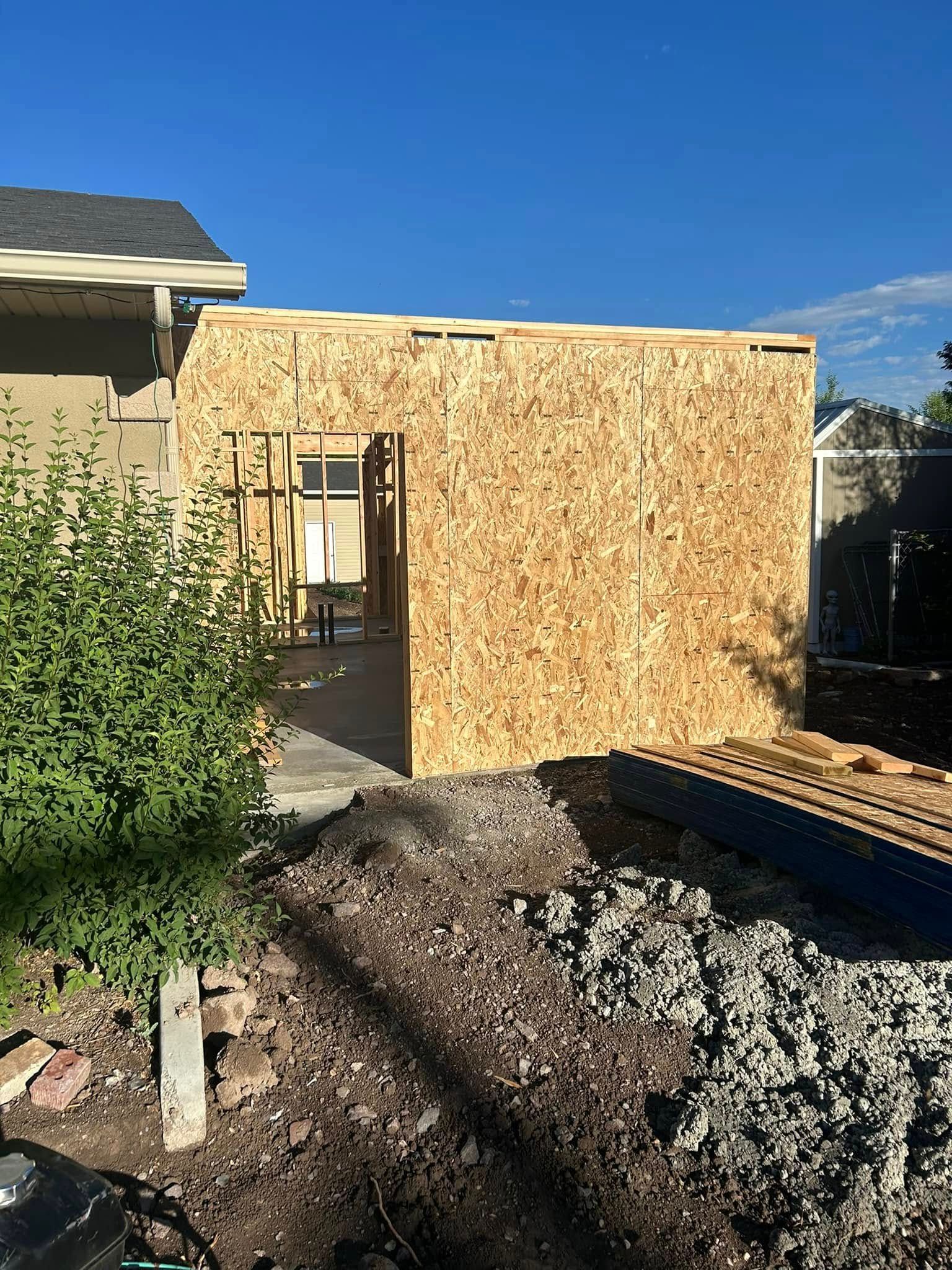 An outdoor construction site showing a wooden frame addition attached to an existing house under a clear blue sky.