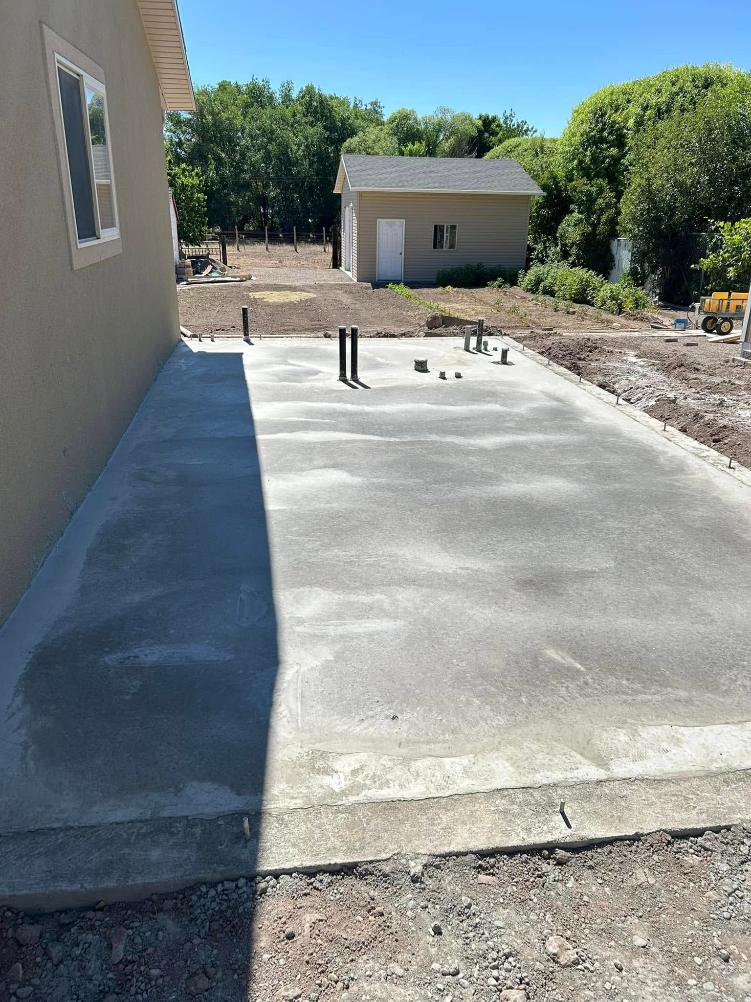 A freshly poured concrete foundation slab sits beside a house, featuring several protruding pipes, with a shed nearby.