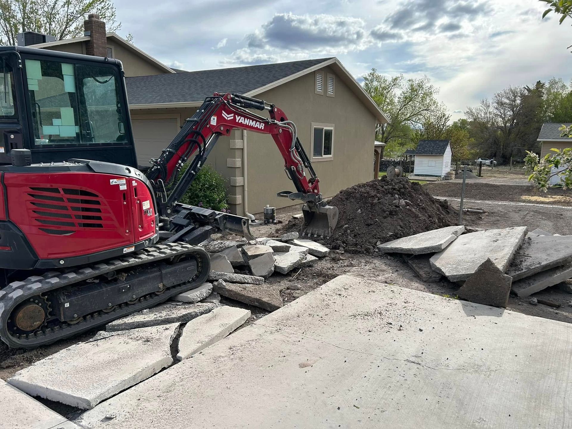 A red excavator sits in a residential yard near a house, breaking up a concrete driveway into a pile of rubble.
