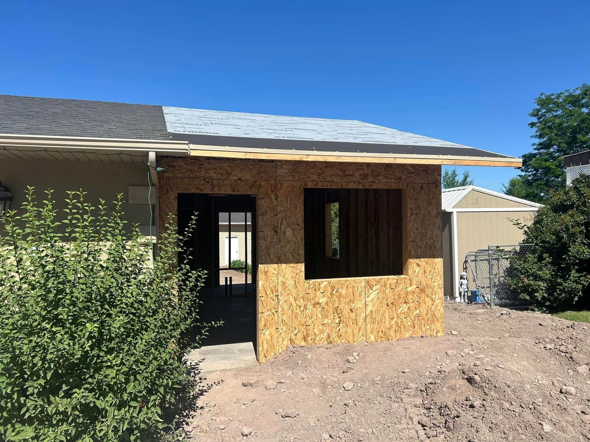 A house addition under construction with exposed wooden framing and OSB panels, featuring a partially roofed entryway.