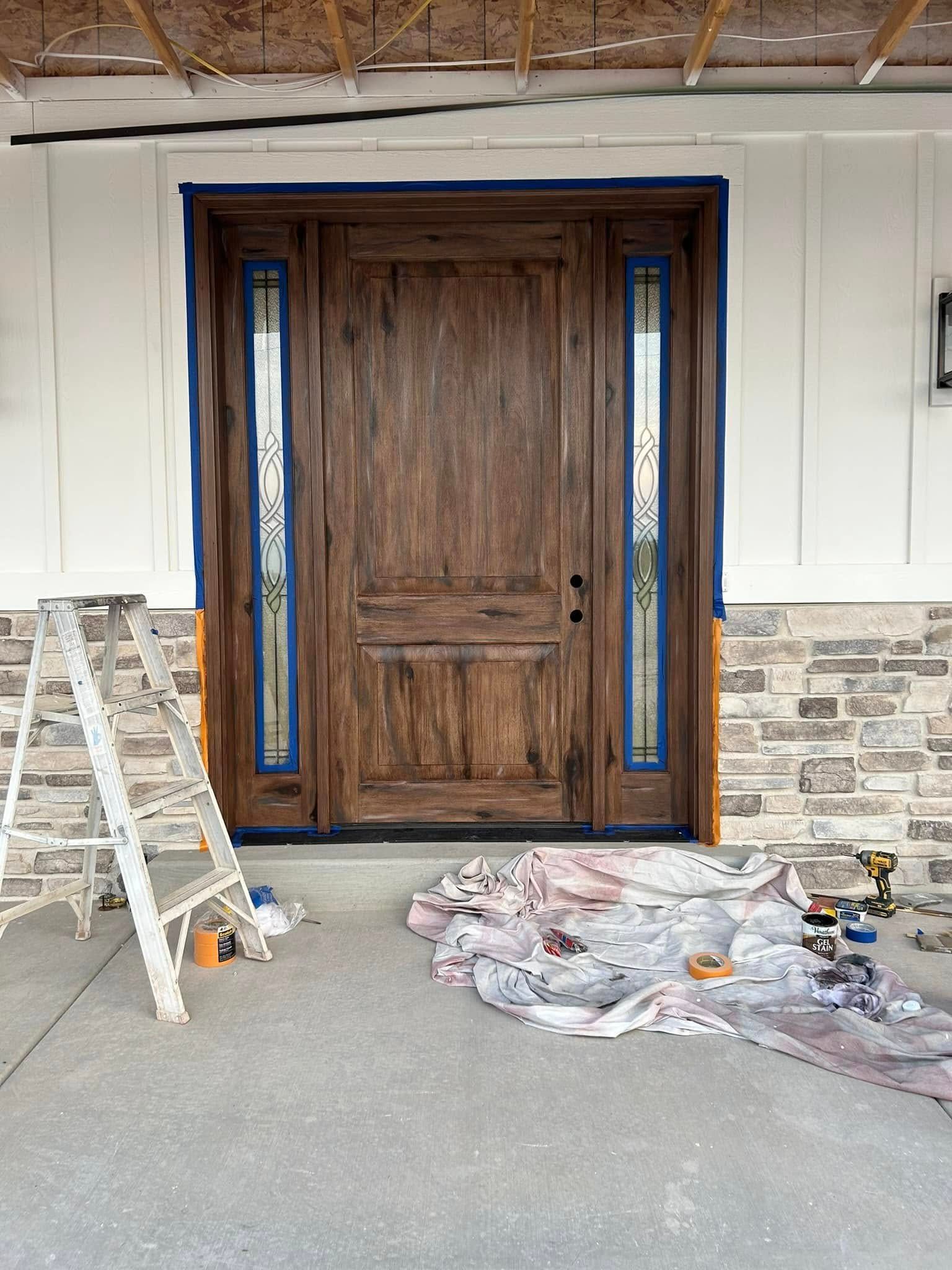 A dark wood front door with glass sidelights is being prepped for painting, with blue tape applied and a ladder nearby.