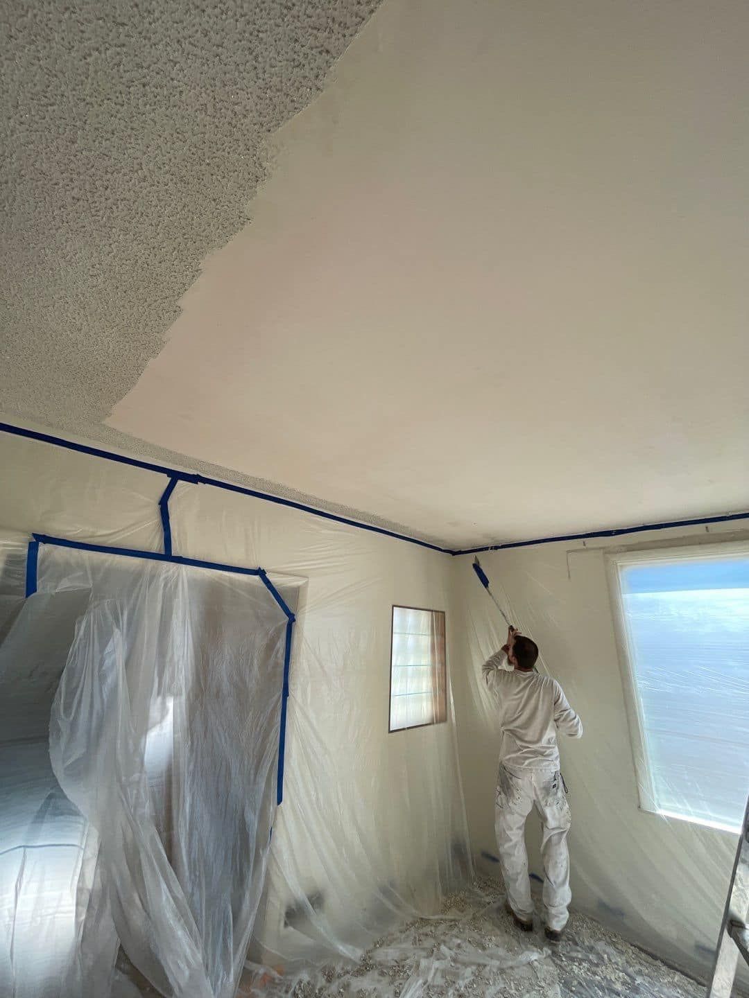 A person in white protective coveralls uses a long-handled roller to paint a ceiling, transitioning from popcorn to smooth.