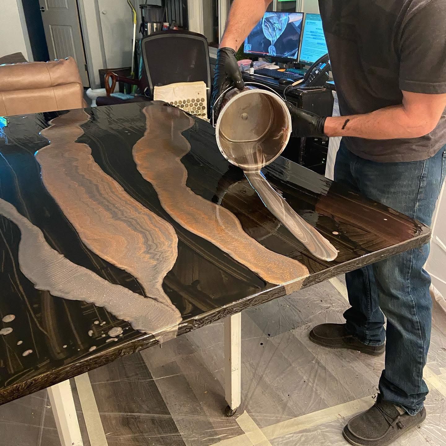 A person wearing black gloves pours shimmering, light brown epoxy onto a dark, glossy table with existing resin channels.