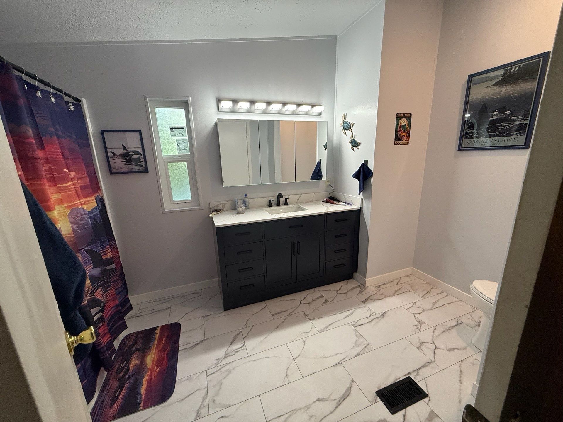 A bathroom with white marbled tile flooring, a dark vanity, a light-colored countertop, and a shower with a colorful curtain.