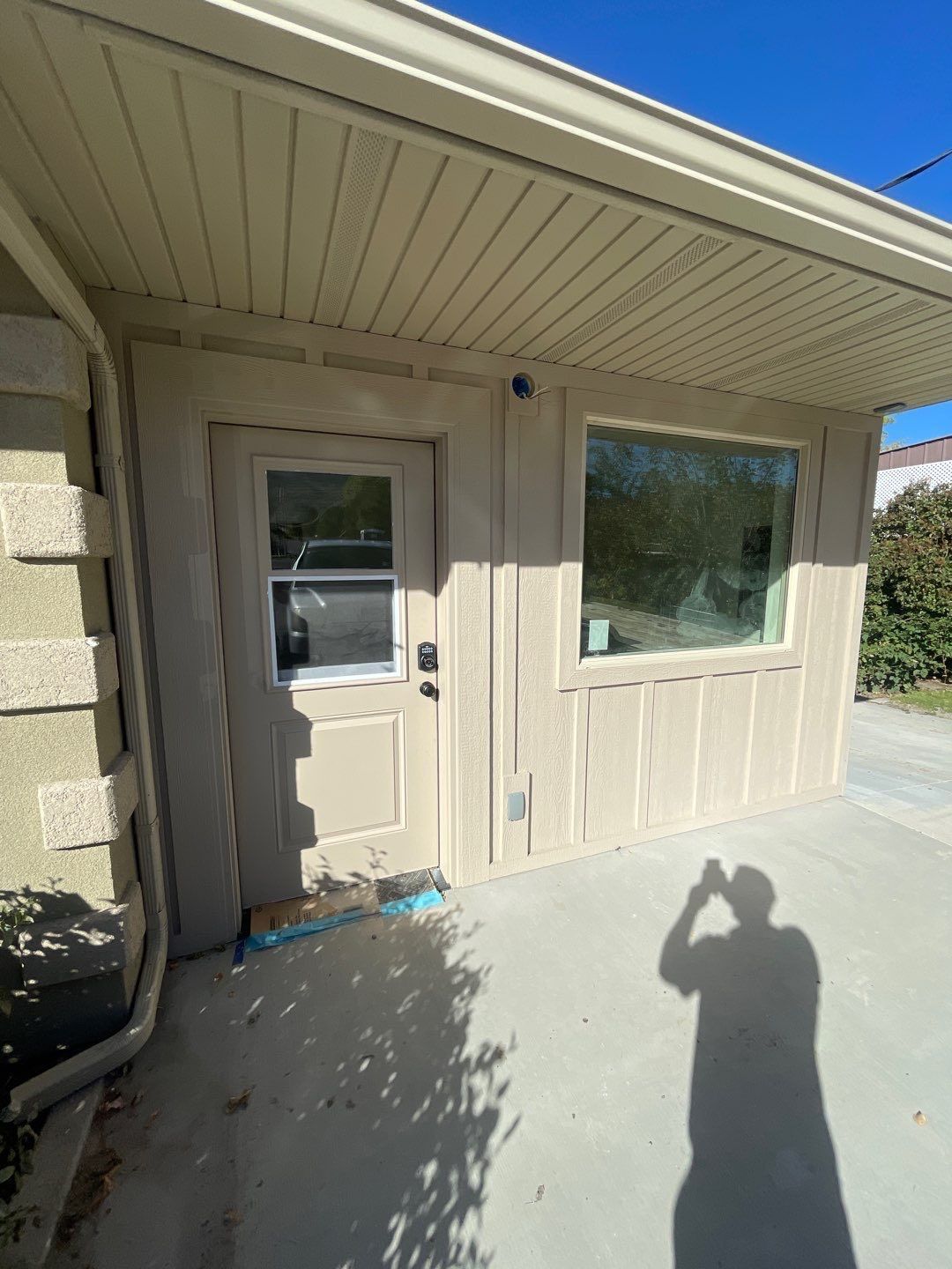 A beige exterior wall with a door and a window under an overhanging roof, with a person’s shadow cast on the concrete.