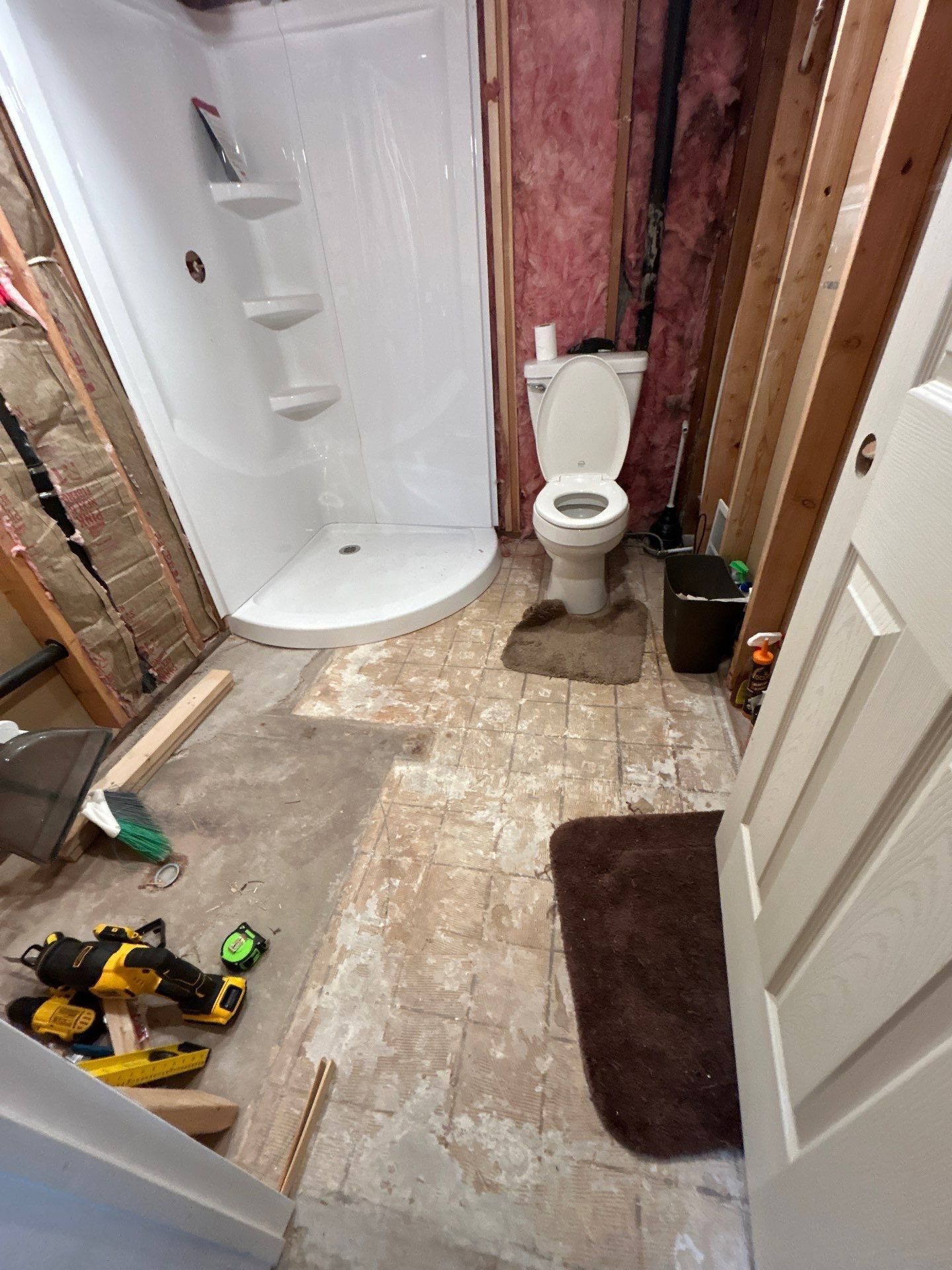 An unfinished bathroom renovation with a white shower stall, a toilet, exposed insulation in the walls, and tools on floor.