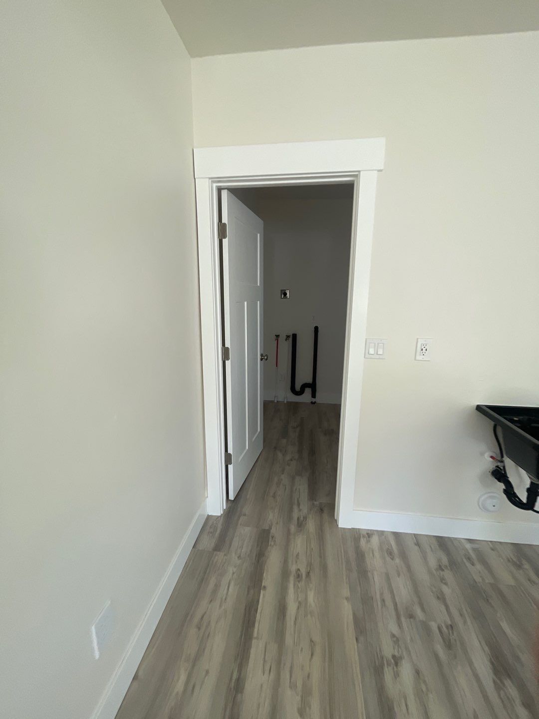 An open doorway leads to a small utility room with exposed plumbing pipes against the back wall, viewed from a room.