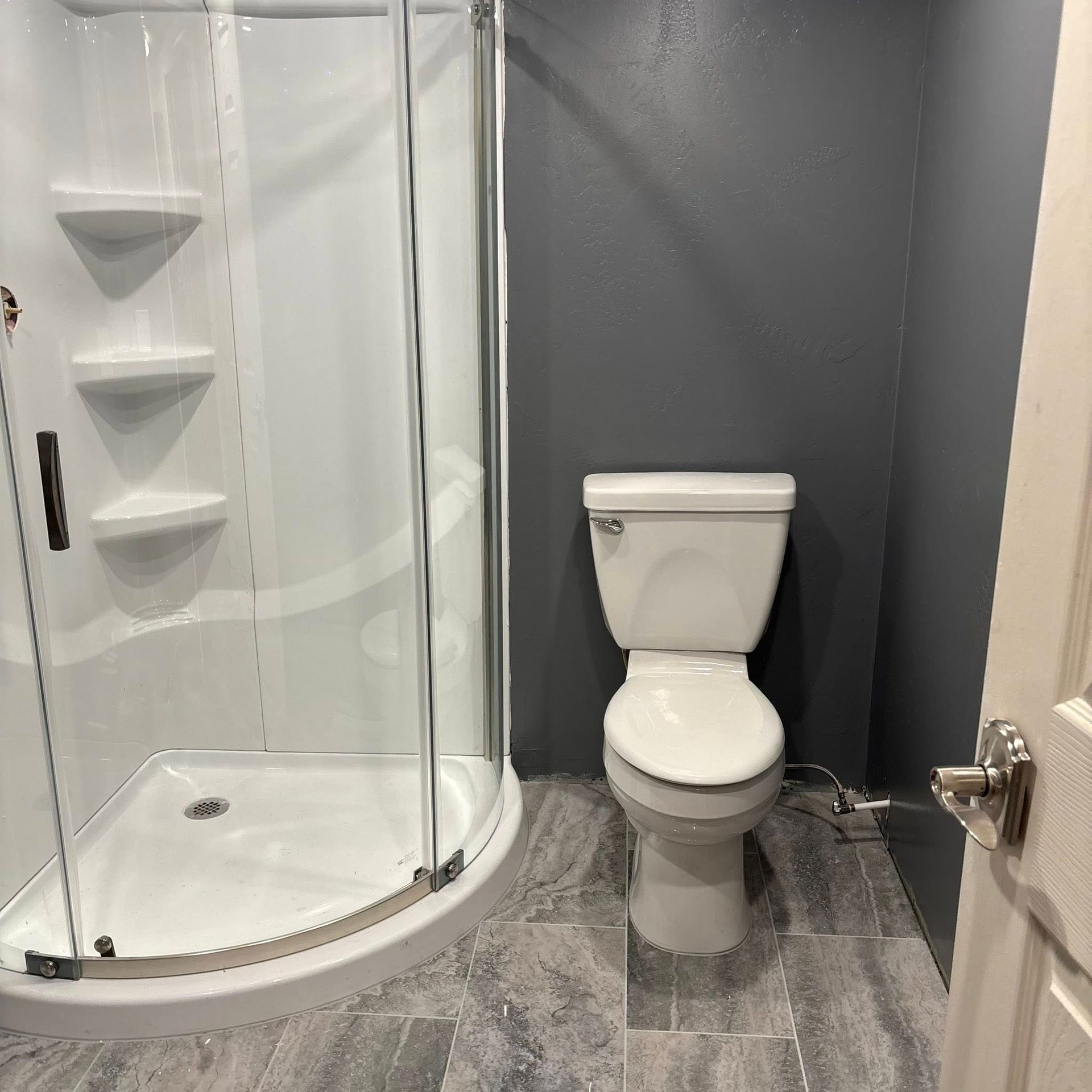 A bathroom with a corner shower stall, a white toilet against a dark gray wall, and gray marbled tile flooring.