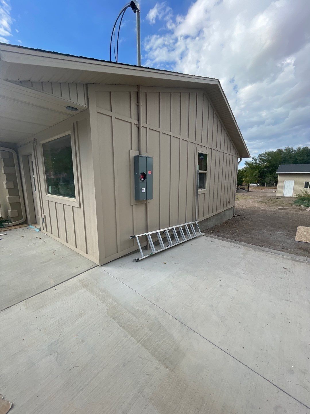 Tan house exterior featuring a grey electrical box, a roof mast, and a metal ladder lying on the concrete patio.