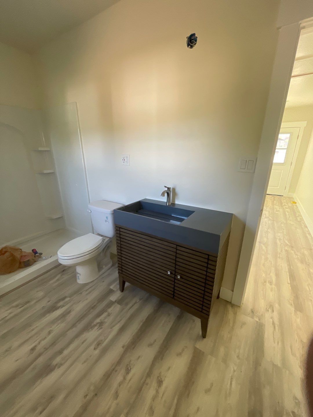Bathroom with a wood-slatted vanity, gray sink, toilet, white shower stall, and light wood-patterned flooring.