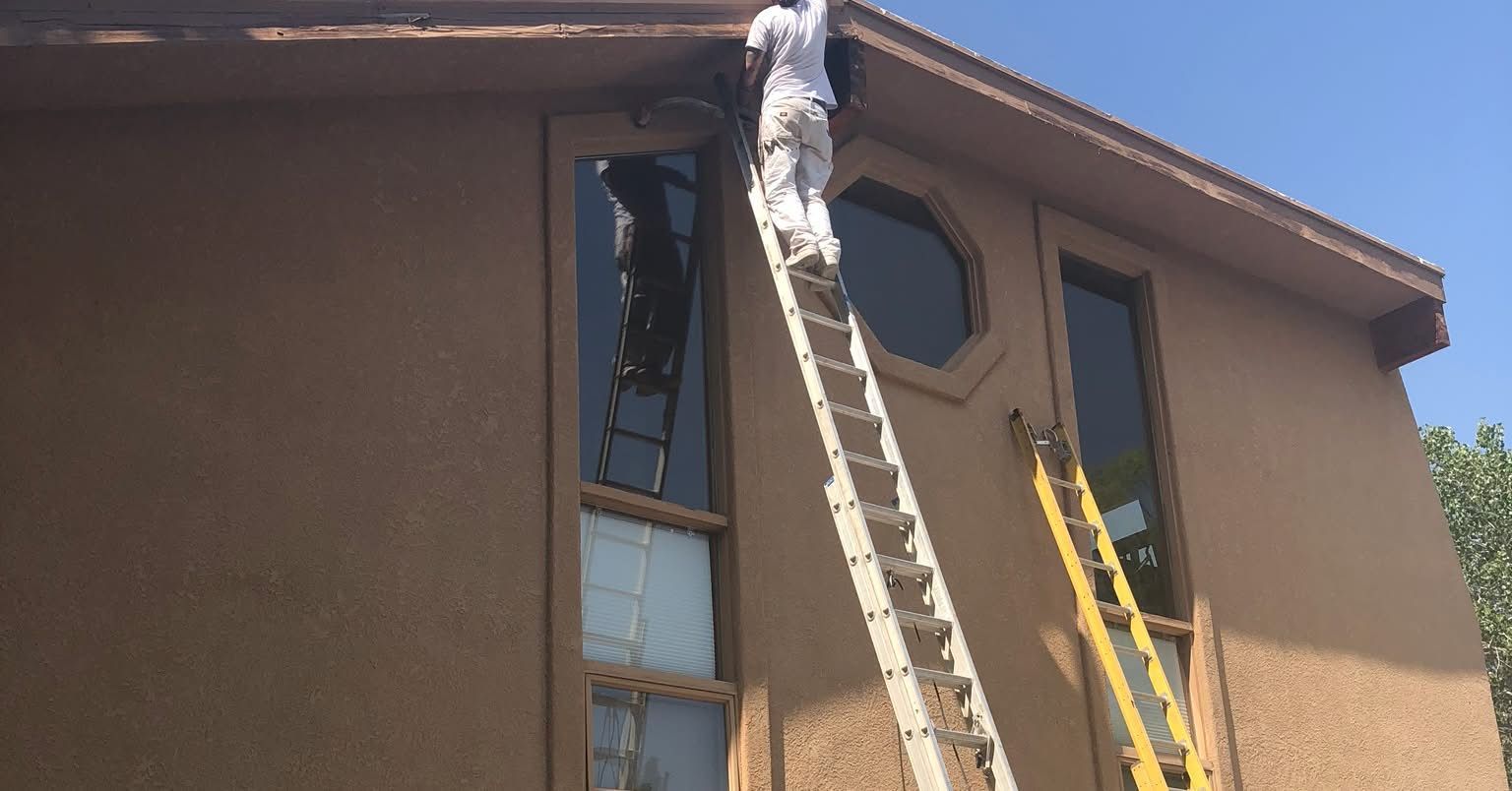 A person in white clothing climbs a tall ladder leaning against the tan, stucco-textured wall of a multi-story house.